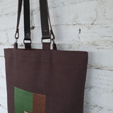Brown tote bag with green and brown square design against a white brick wall.
