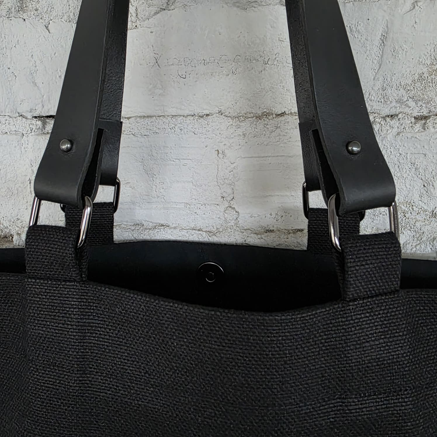 Close-up of a black linen-cotton tote bag showing the removable genuine leather handles, metal rings, and sturdy stitching, highlighting quality