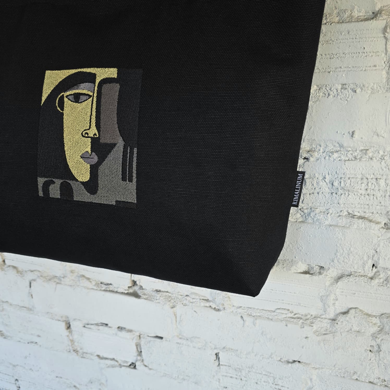 A black canvas tote bag featuring a stylized cubist face graphic in gold, gray, and black, hanging against a white brick wall. A small side tag displays the brand 'RIMALINUM'.