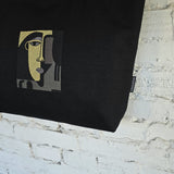 A black canvas tote bag featuring a stylized cubist face graphic in gold, gray, and black, hanging against a white brick wall. A small side tag displays the brand 'RIMALINUM'.