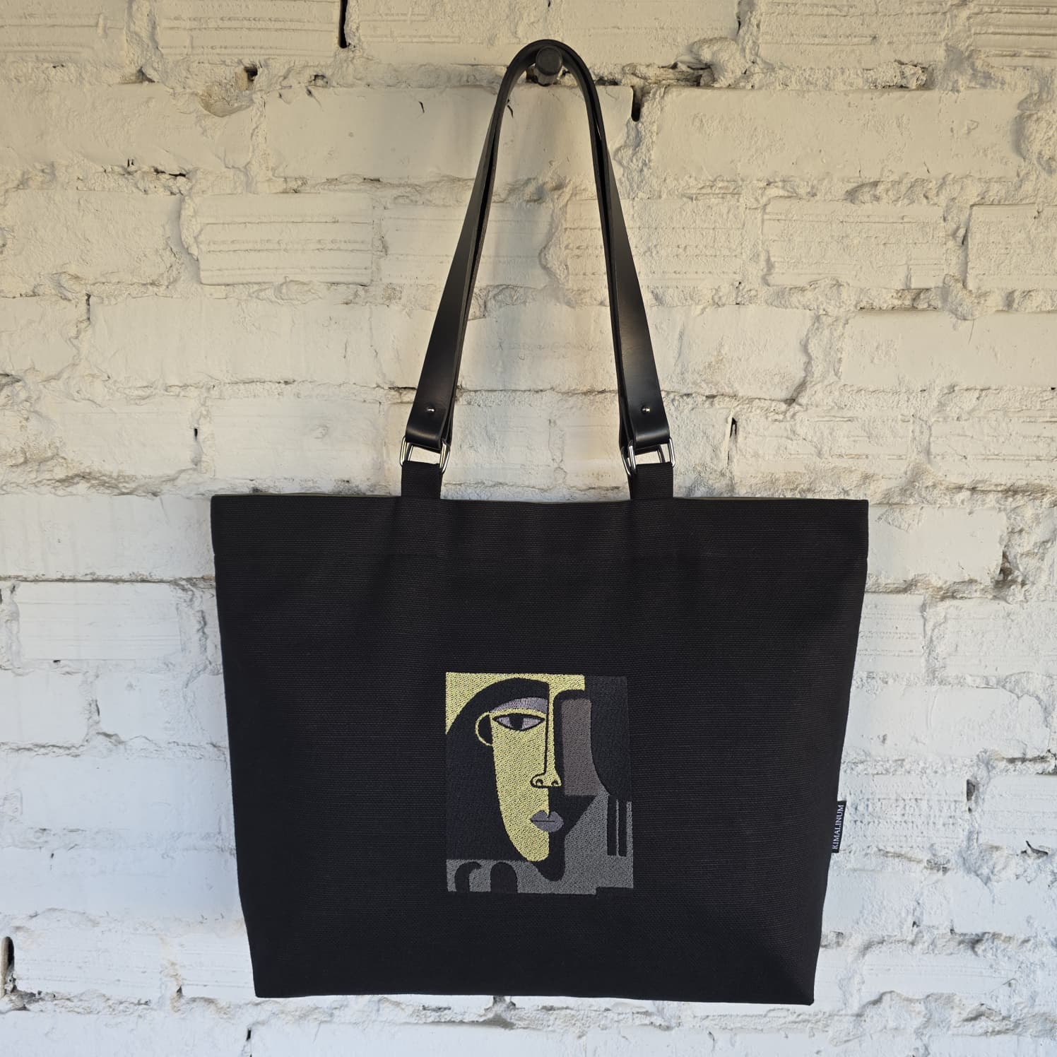A black canvas tote bag with black shoulder straps, featuring a stylized cubist face graphic in olive green, gray, and beige on the front, hanging against a white brick wall.