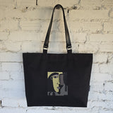 A black canvas tote bag with black shoulder straps, featuring a stylized cubist face graphic in olive green, gray, and beige on the front, hanging against a white brick wall.