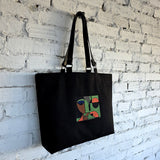 Black tote bag with a colorful abstract design on a white brick wall