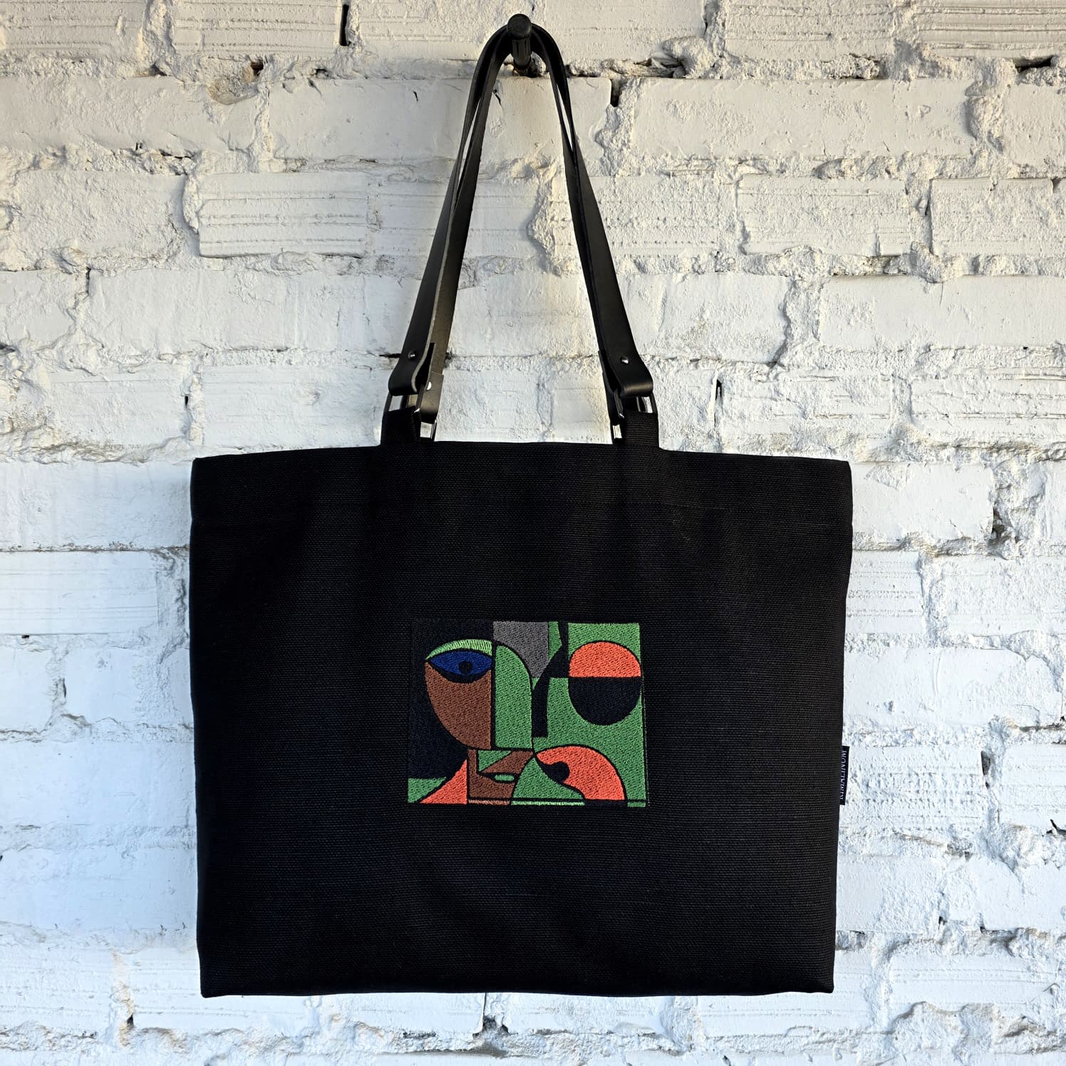 Front view of black canvas tote bag with embroidered abstract face artwork