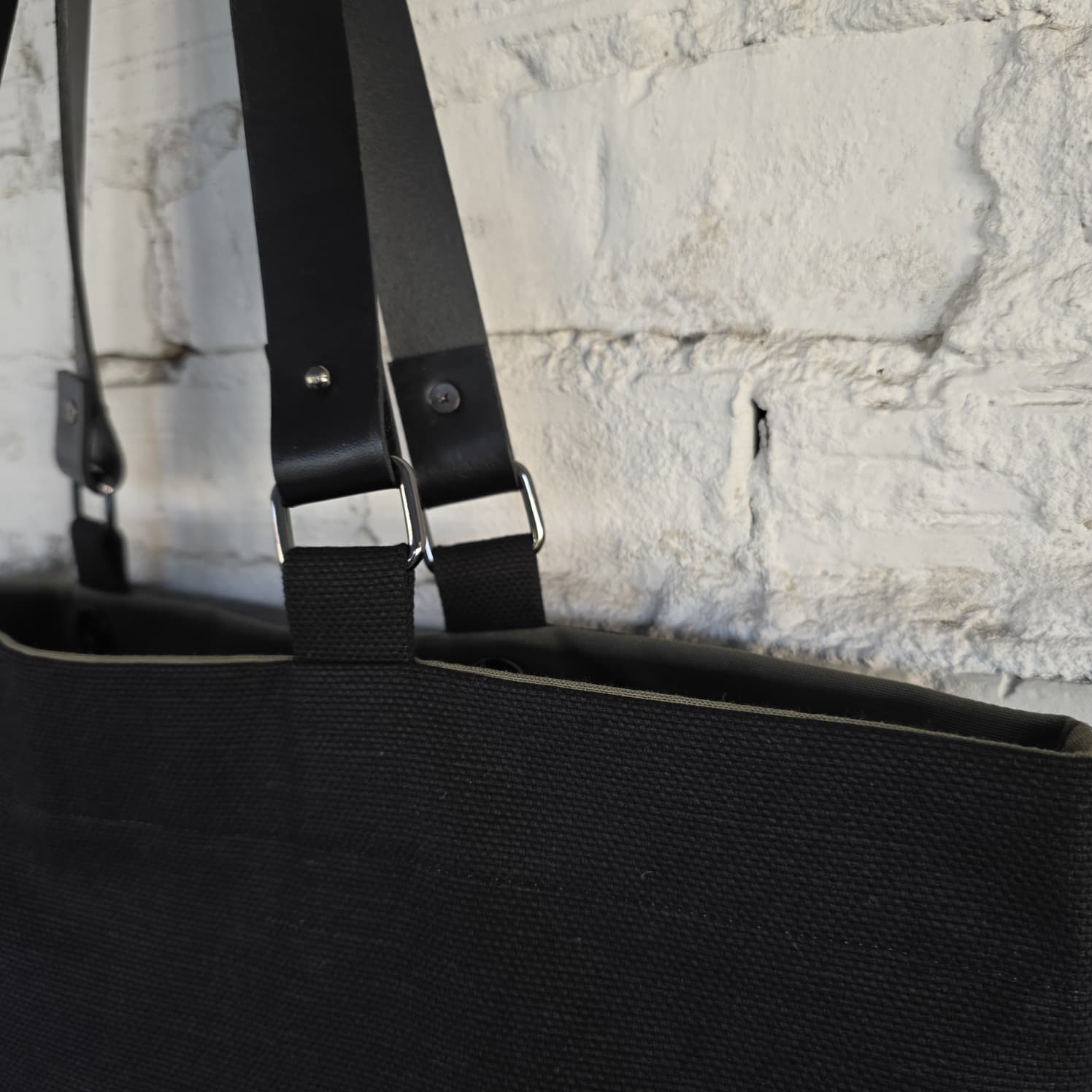A close-up of a black canvas tote bag with black straps and silver metal buckles, shown against a white painted brick wall.