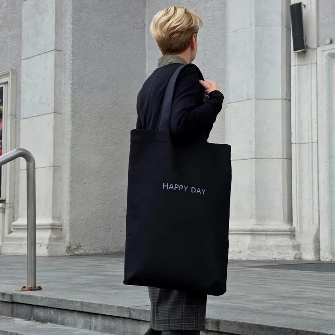 Person holding a black tote bag with 'HAPPY DAY' text in front of a gray building.