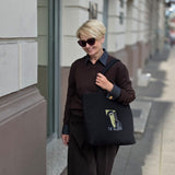 A woman walking on a city sidewalk wearing a dark brown long-sleeve shirt and matching wide-leg trousers, carrying a black tote bag with a graphic print. She is wearing sunglasses and dark sneakers.