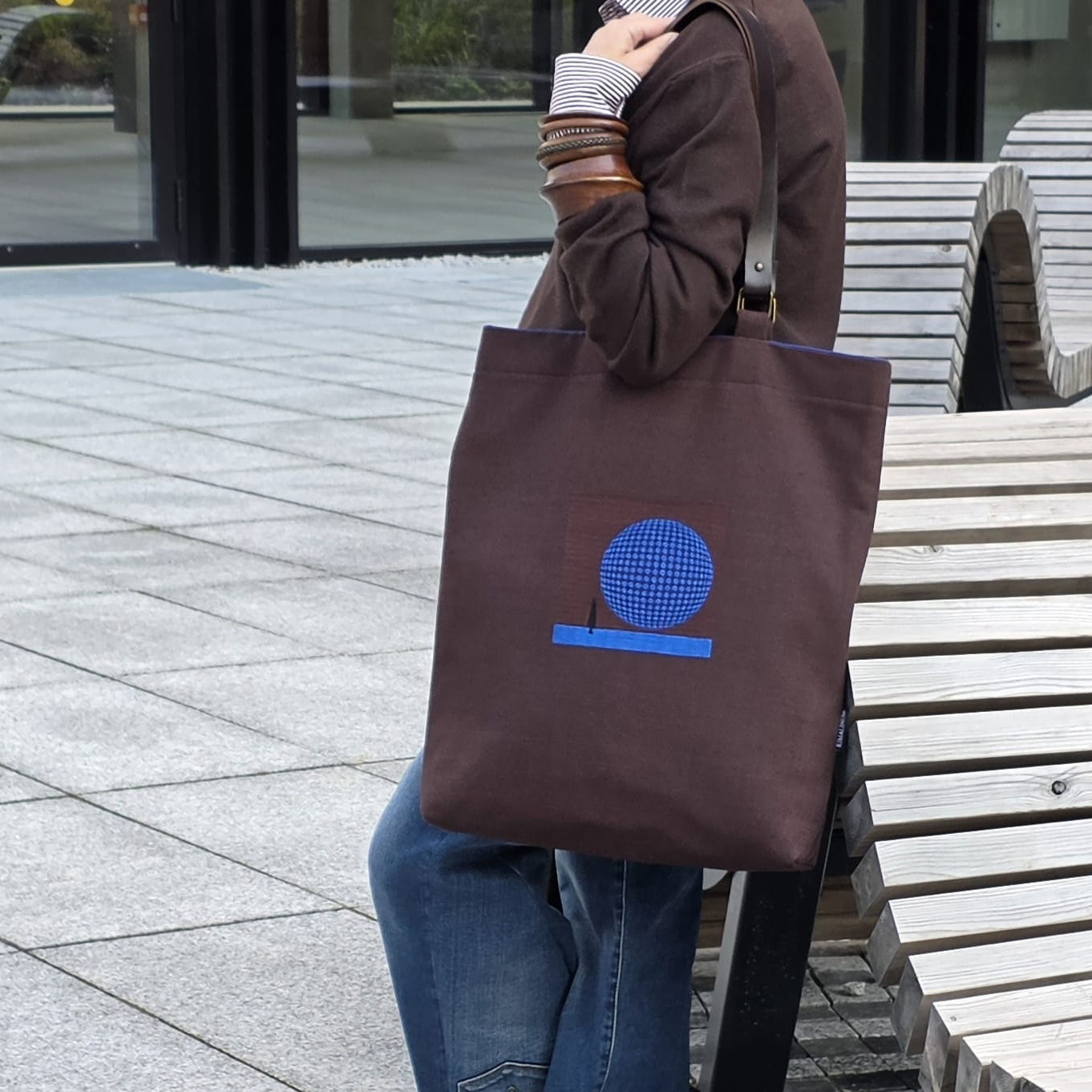 A person sitting outdoors next to a bench carrying a dark brown canvas tote bag with a blue embroidered design the front.