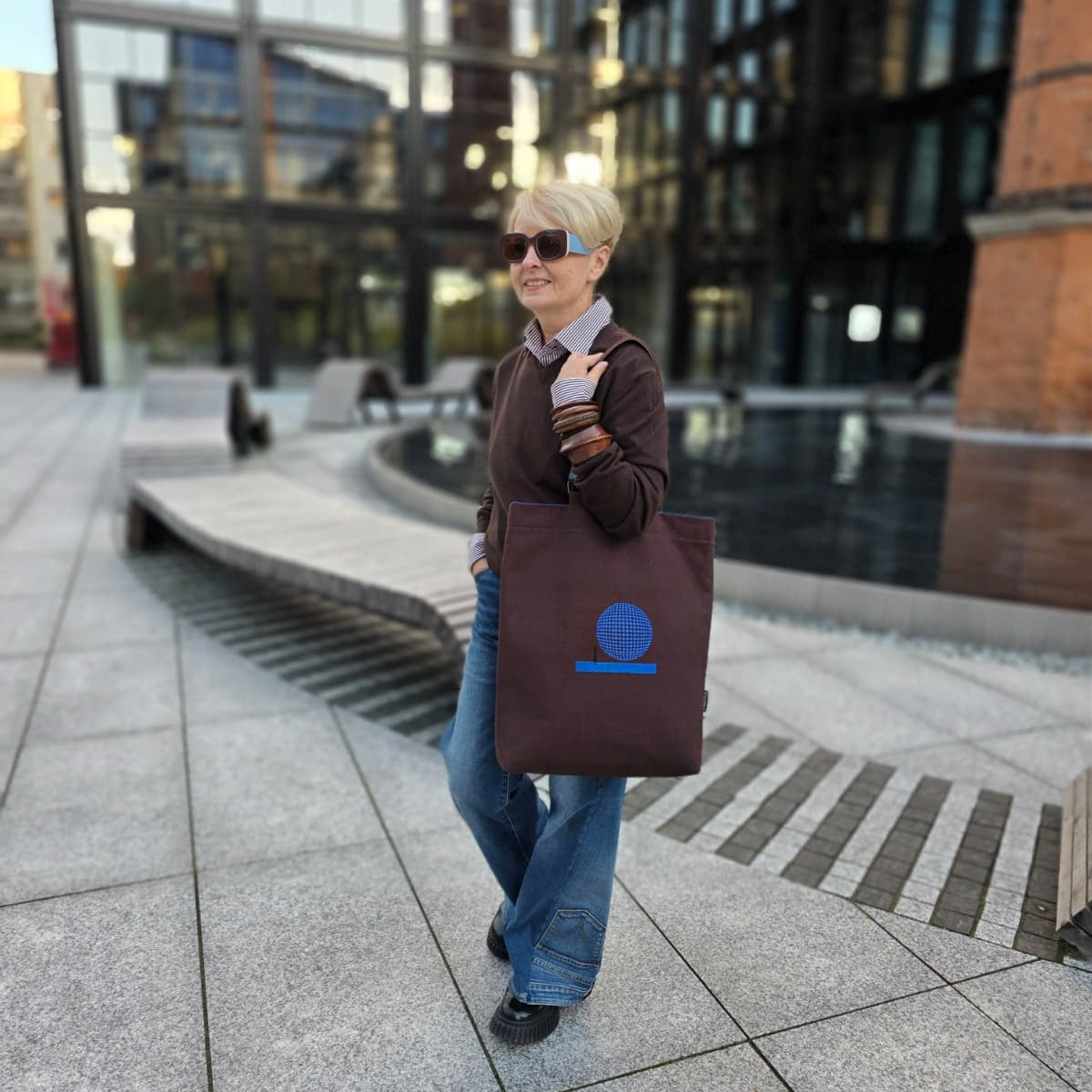 A woman with short blonde hair and sunglasses walks outdoors carrying a dark brown tote bag with a blue  embroidered design. The background shows a modern glass building and paved walkway.