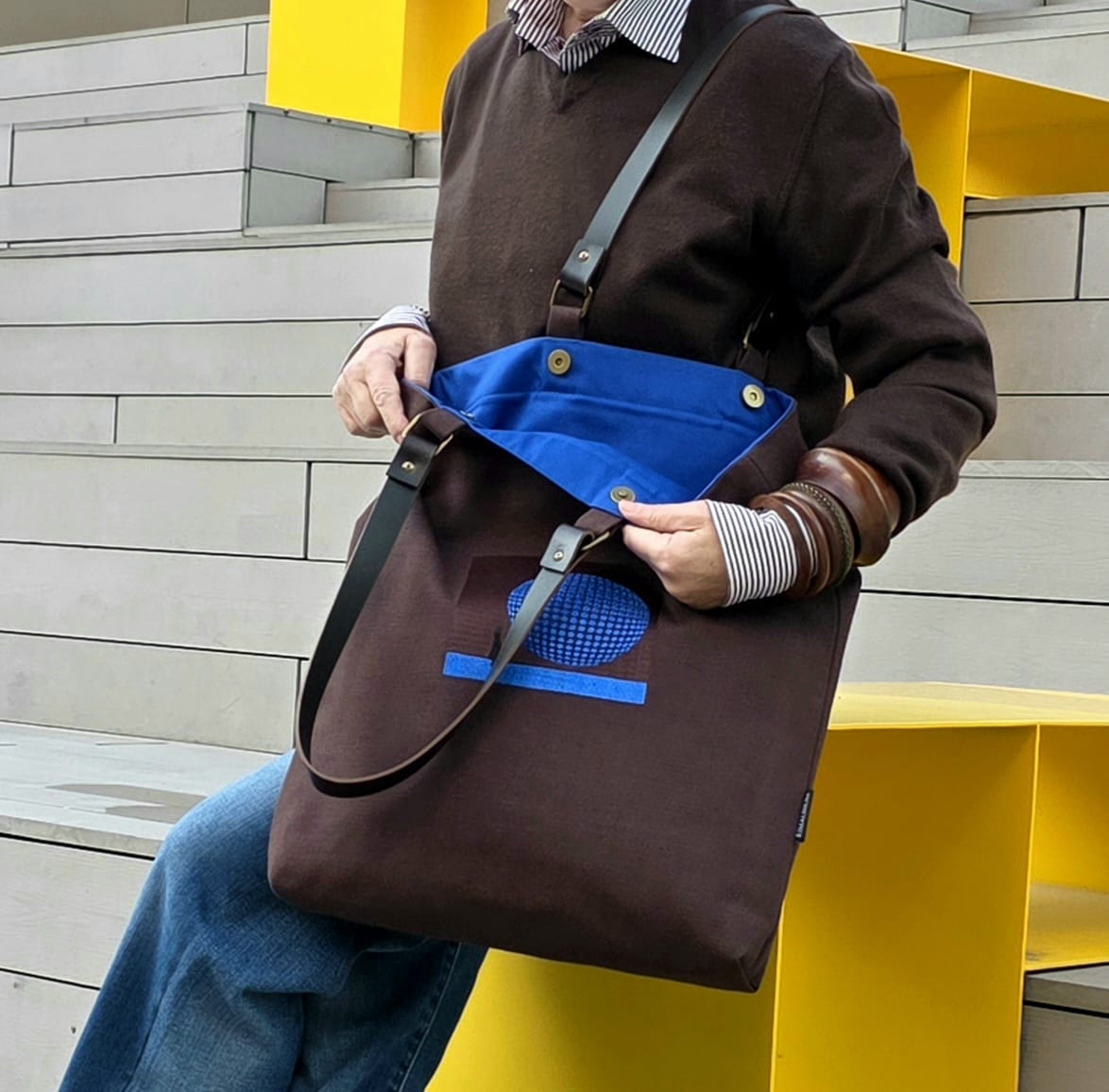 A person wearing a brown sweater and blue jeans carries a large brown tote bag with a bright blue interior and blue graphic detail on the front, set against a modern yellow and gray architectural background.