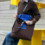 A person wearing a brown sweater and blue jeans carries a large brown tote bag with a bright blue interior and blue graphic detail on the front, set against a modern yellow and gray architectural background.