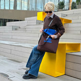 A woman with short blonde hair and sunglasses sits on a modern yellow bench on outdoor concrete steps, holding a brown tote bag . She is wearing a brown jacket, blue wide-leg jeans, and black shoes, with a contemporary glass building in the background.