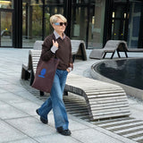 A woman with short blonde hair and sunglasses walks across a modern outdoor plaza carrying a brown tote bag with a blue logo, wearing a brown jacket, white shirt, and blue jeans.
