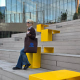 A woman sits on outdoor concrete steps beside bright yellow modern seating, holding a brown tote bag with a blue embroidered design. A glass building is visible in the background.