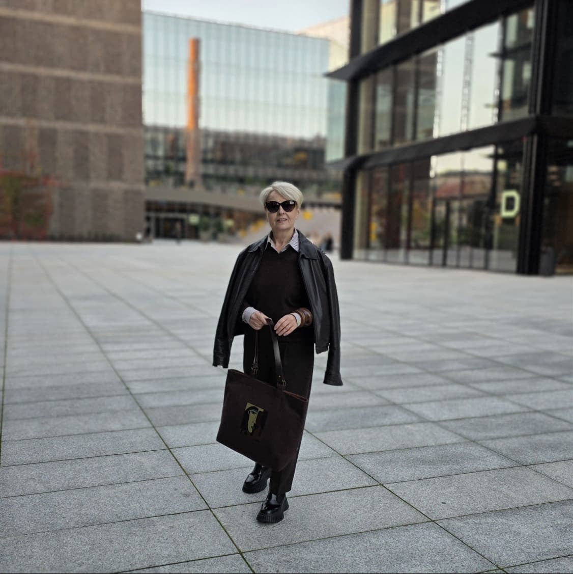 A person walking outdoors in a modern plaza, wearing a black outfit with a black leather jacket, black sunglasses, and carrying a dark brown leather briefcase.