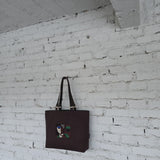 Large brown embroidered tote bag with leather handles displayed on white textured brick wall