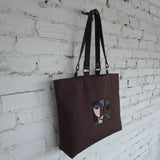 Brown canvas tote bag with embroidered abstract art design and leather shoulder handles hanging against white brick wall