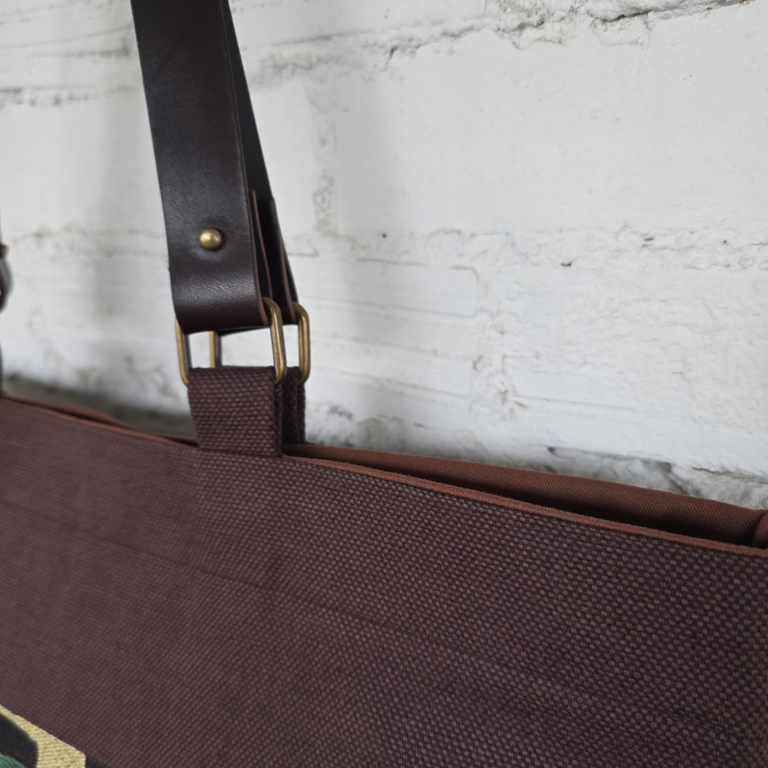 Close-up of a dark brown canvas tote bag with a dark brown leather shoulder strap and brass buckle, set against a white brick wall background.
