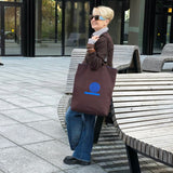 A woman with short blonde hair and sunglasses stands on a paved outdoor plaza next to wooden benches, holding a dark brown tote bag with a blue embroidered design.