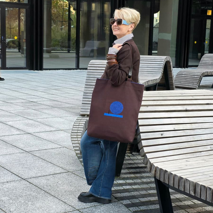 A woman with short blonde hair and sunglasses stands on a paved outdoor plaza next to wooden benches, holding a dark brown tote bag with a blue embroidered design.