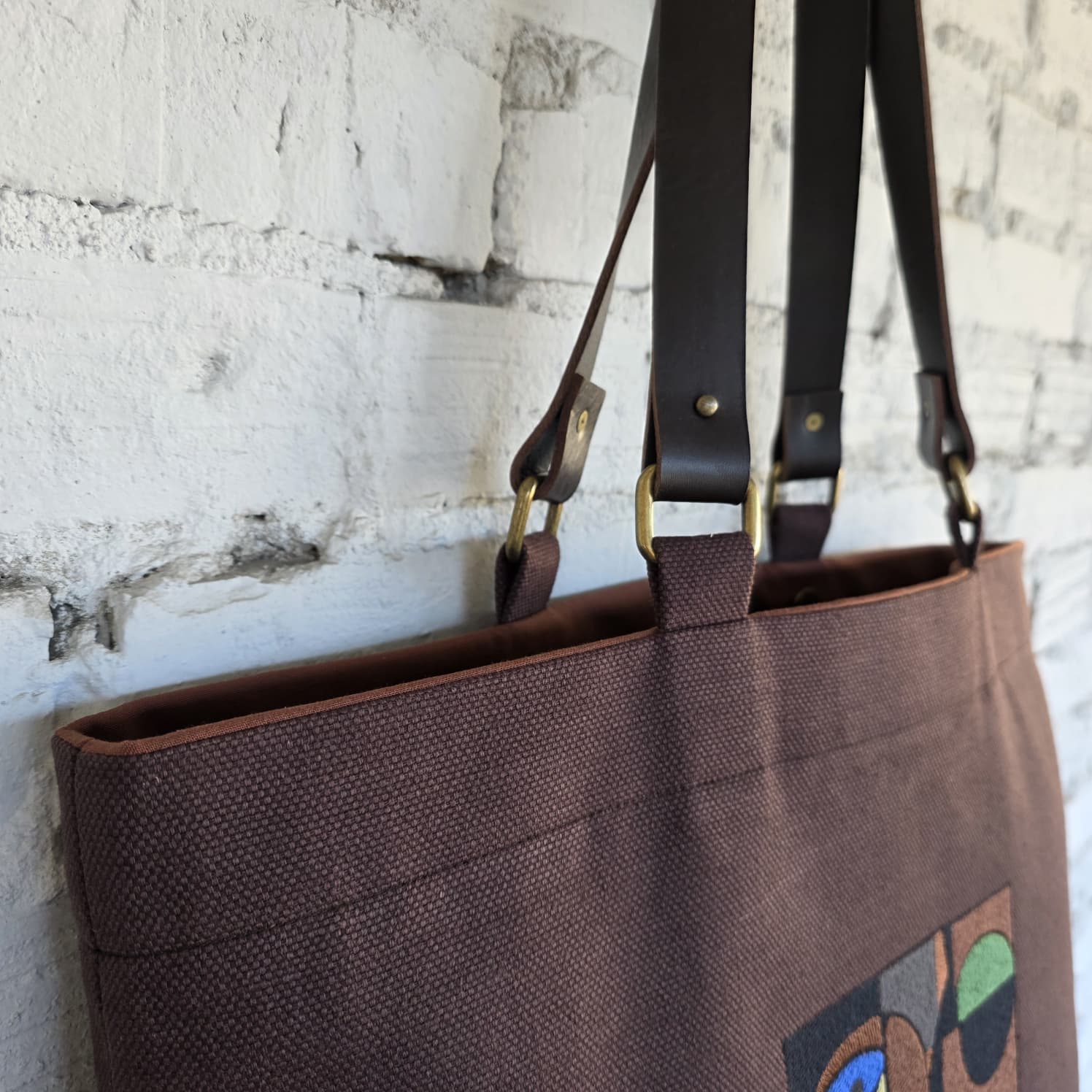 Brown tote bag with decorative design on a textured white wall background
