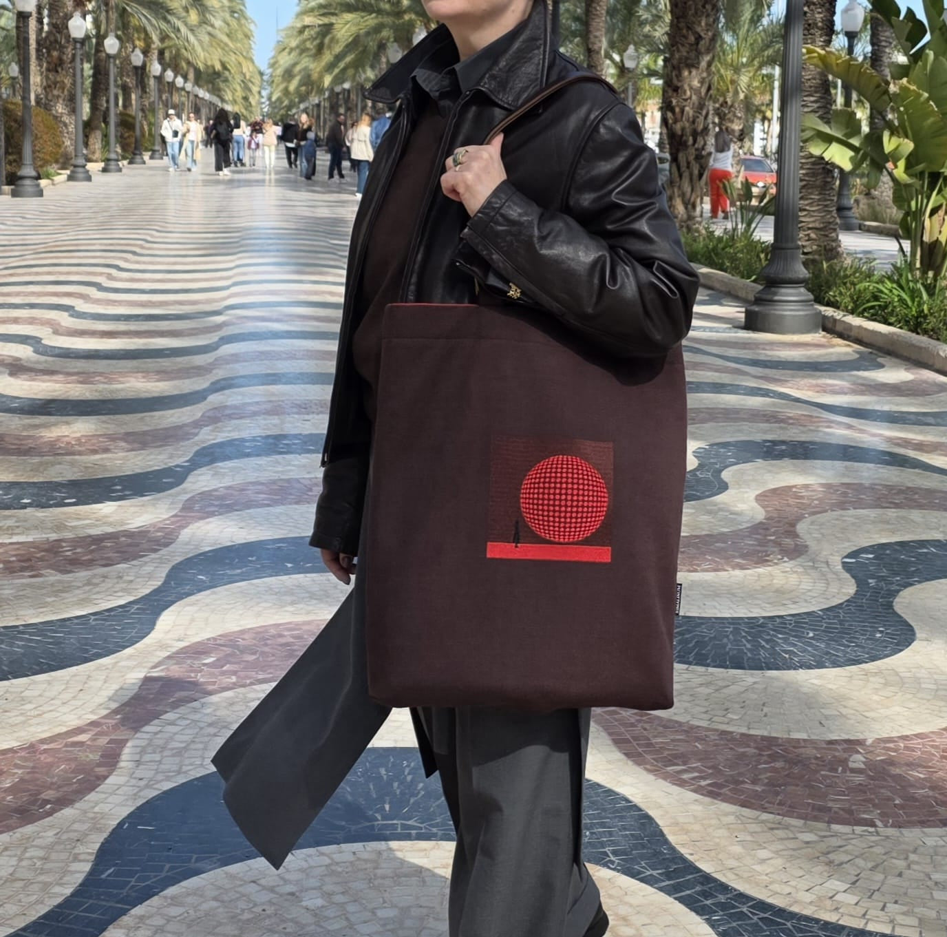 Woman carrying brown tote bag with red embroidery on city promenade, lifestyle shot