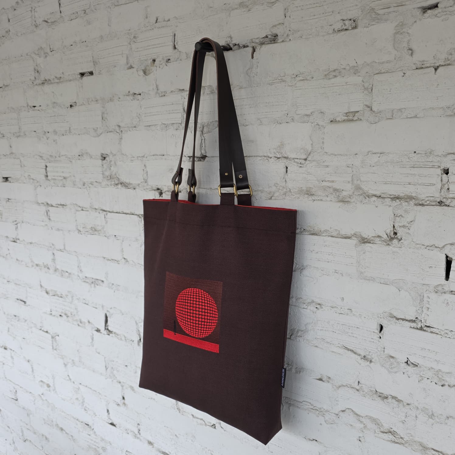 Brown minimalist tote bag with red geometric embroidery hanging against textured wall