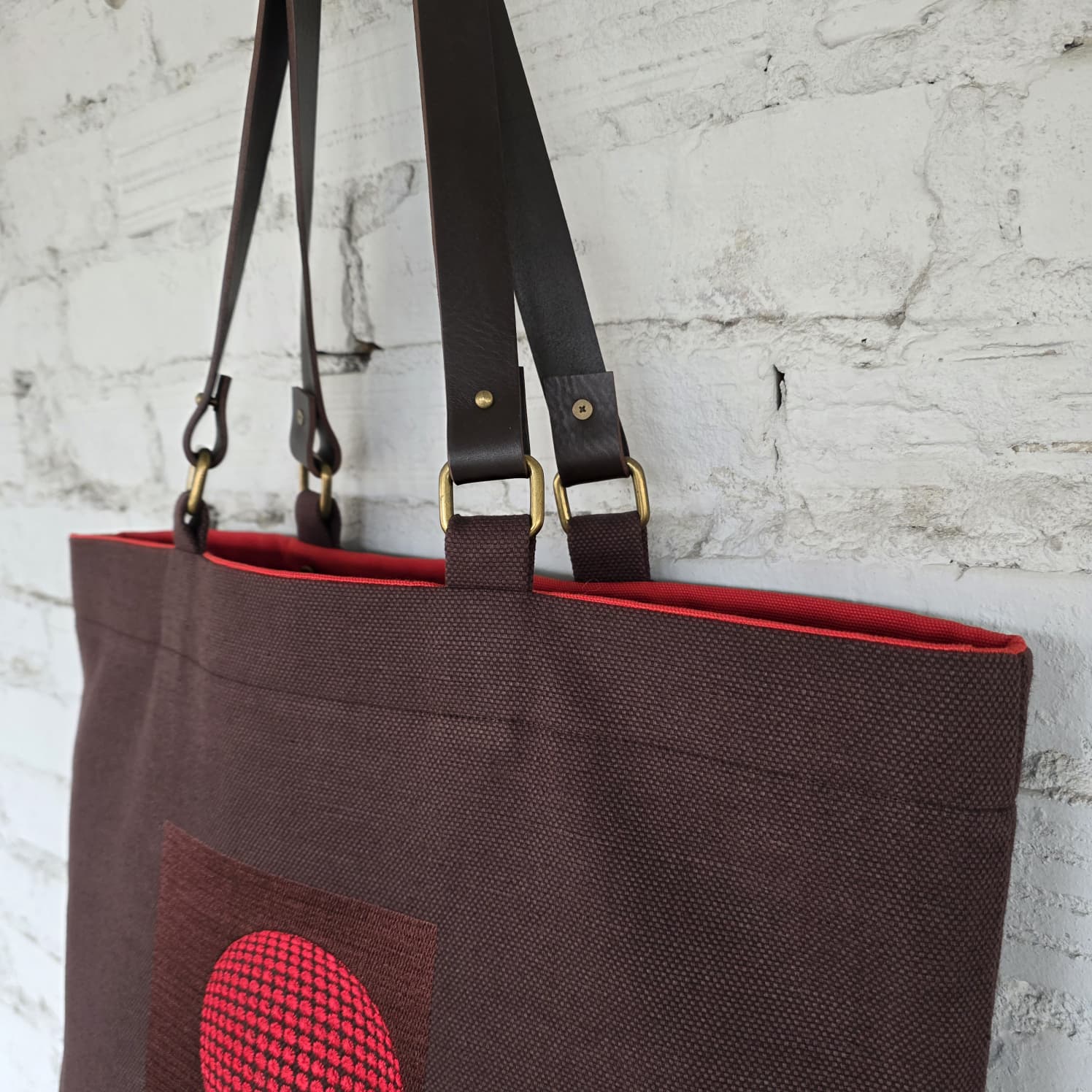 Brown tote bag with red interior and pocket against a textured white wall