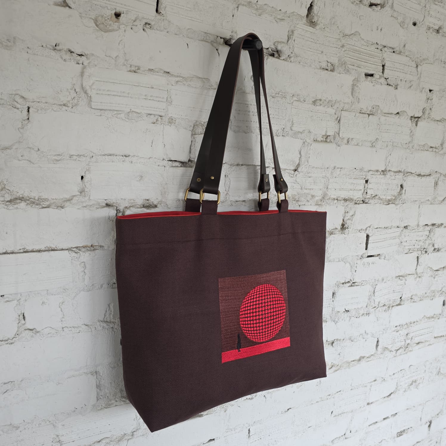 Brown tote bag with red design against a white brick wall
