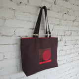 Brown tote bag with red design against a white brick wall
