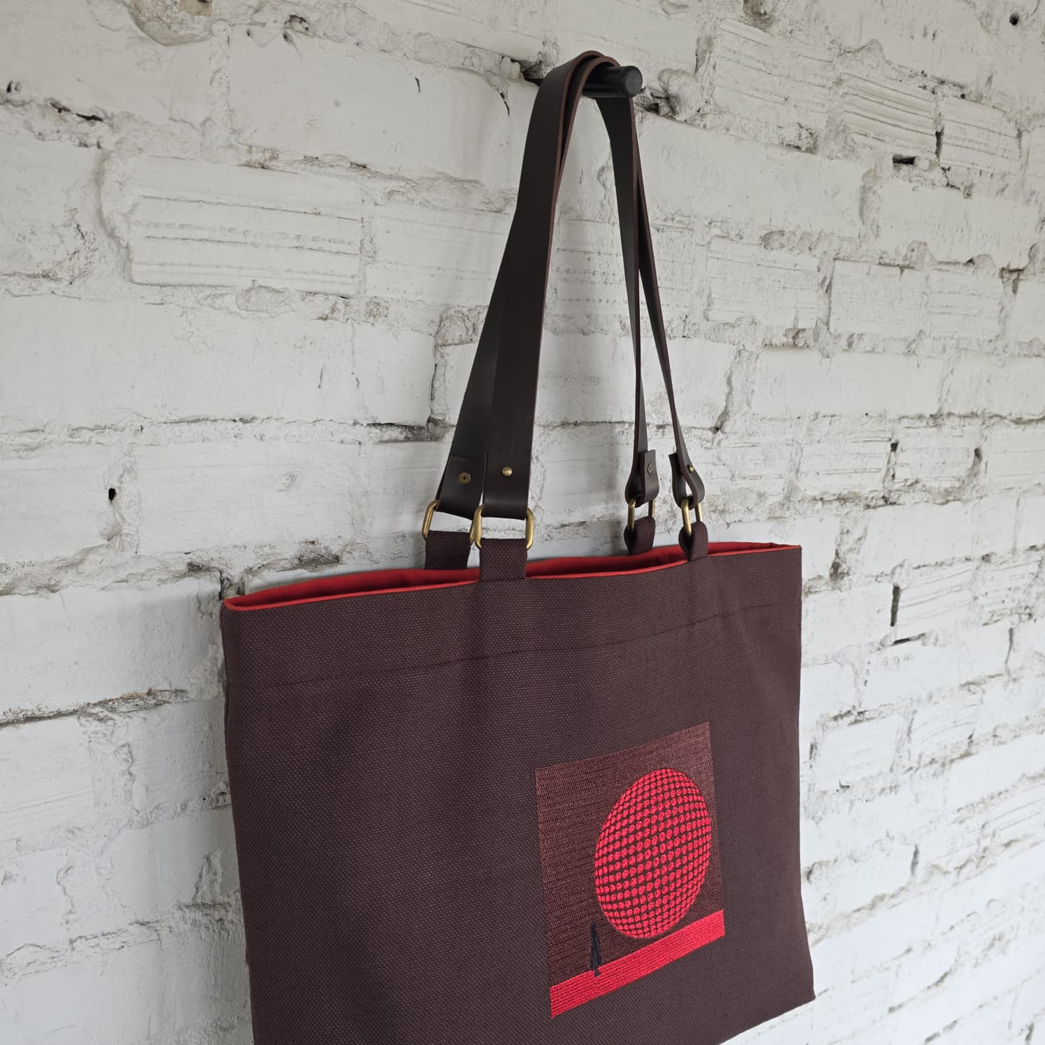Brown linen tote bag with leather handles and red embroidered 3D design hanging on white wall