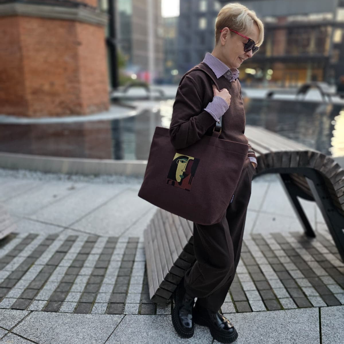 A person standing outdoors on a paved plaza near a brick column and water feature, wearing a dark brown outfit and carrying a large dark brown tote bag with embroiered absract face design