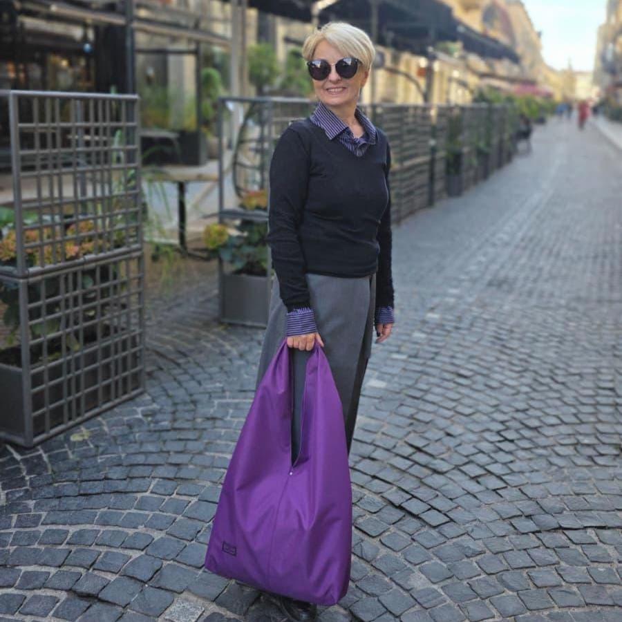 A woman with short blonde hair and sunglasses stands on a cobblestone street, holding a large purple reusable tote bag. She is wearing a black sweater over a purple collared shirt and a gray skirt.