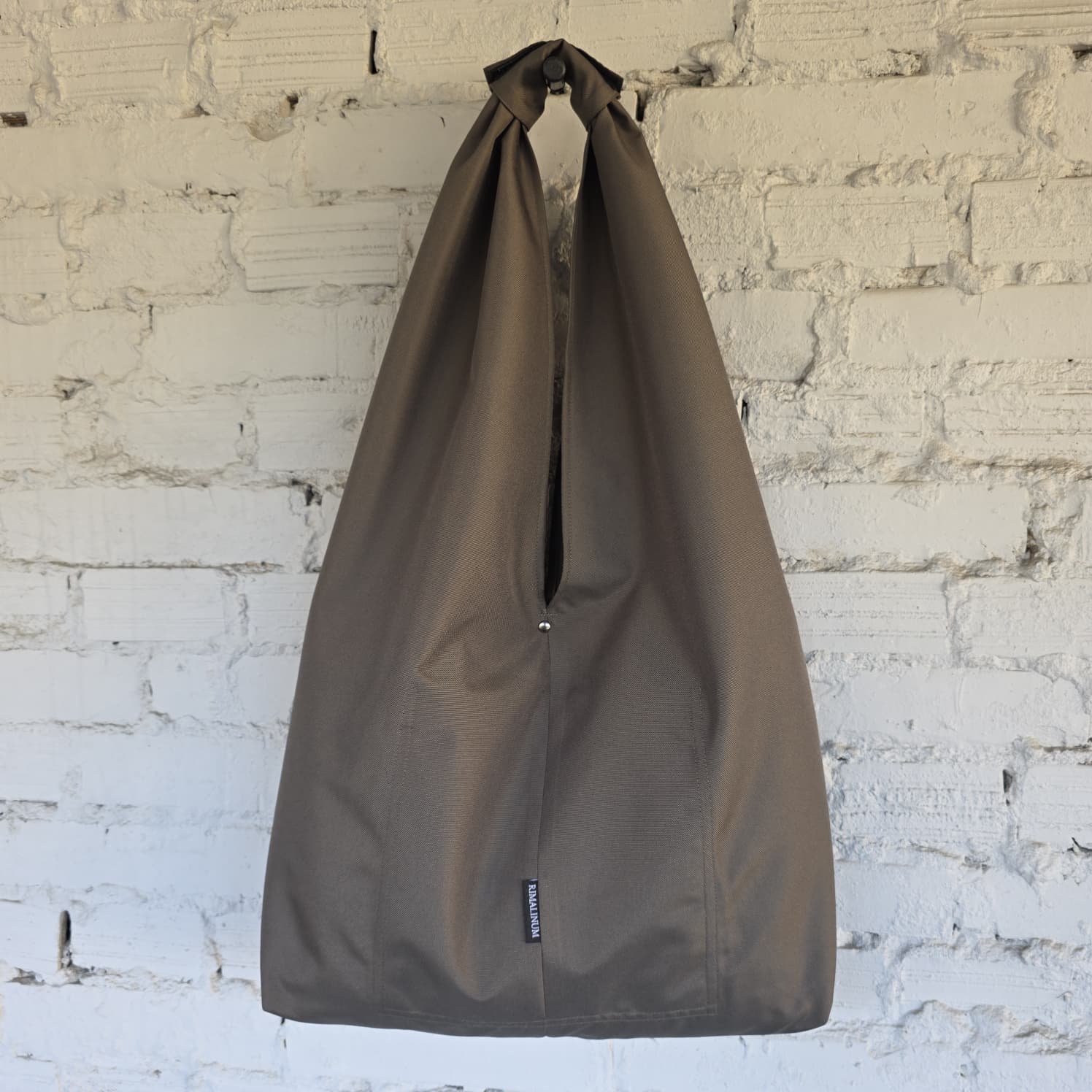 A large khaki waterproof hobo bag with a top loop, hanging against a white brick wall.