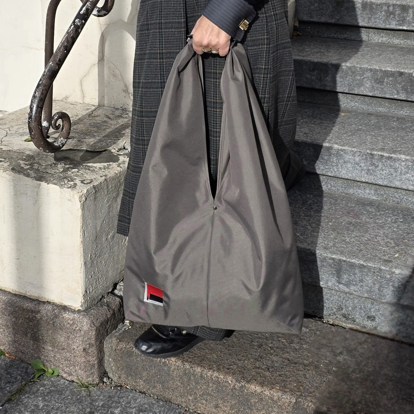 A person standing on stone steps holds a large khaki hobo bag with  a small red, grey, and black embroidered design  near the bottom left corner.