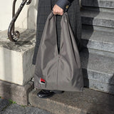 A person standing on stone steps holds a large khaki hobo bag with  a small red, grey, and black embroidered design  near the bottom left corner.
