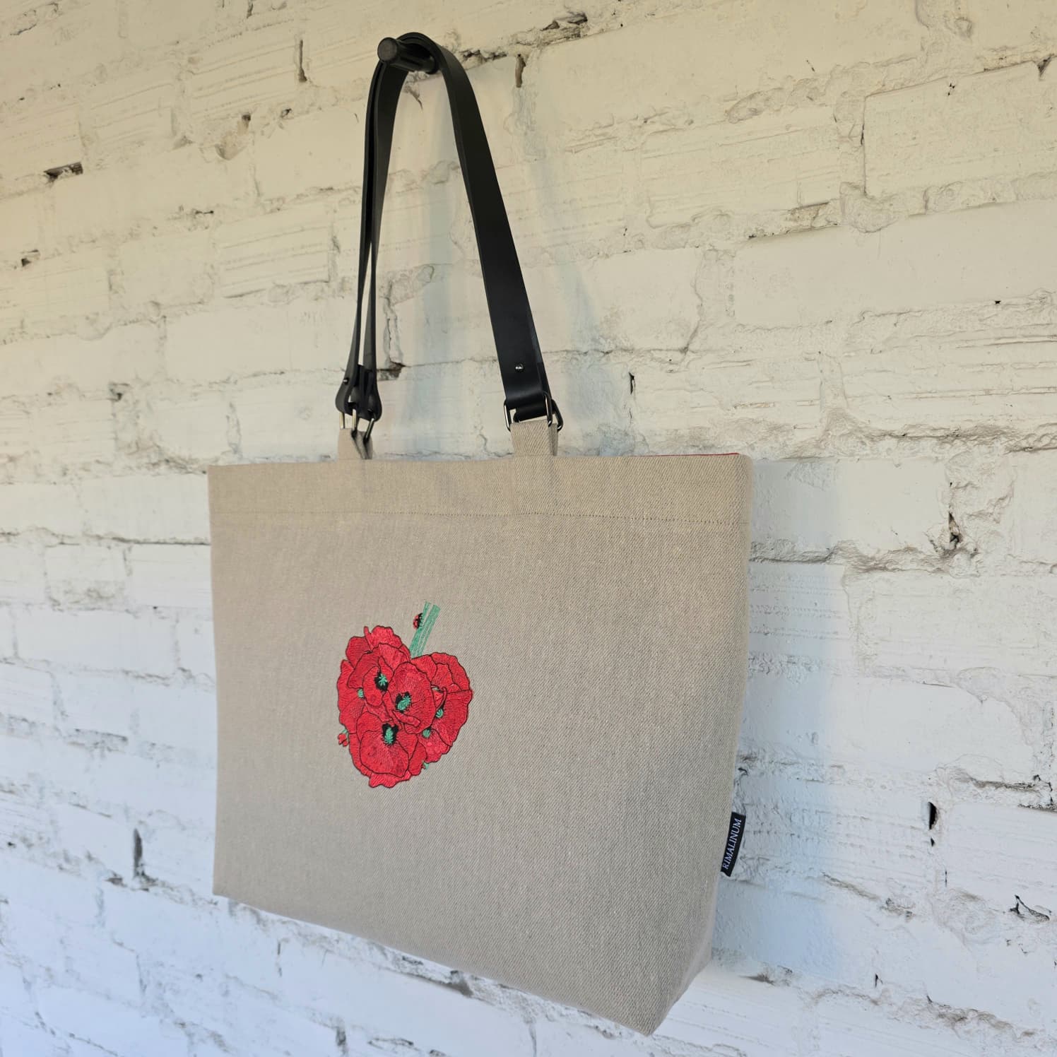 A natural linen tote bag with black shoulder straps, featuring a red embroidered raspberry design on the front, hanging against a white brick wall.