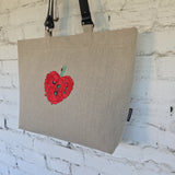 A natural linen tote bag with a black shoulder strap, featuring a red poppy flower graphic on the front, hanging against a white painted brick wall.