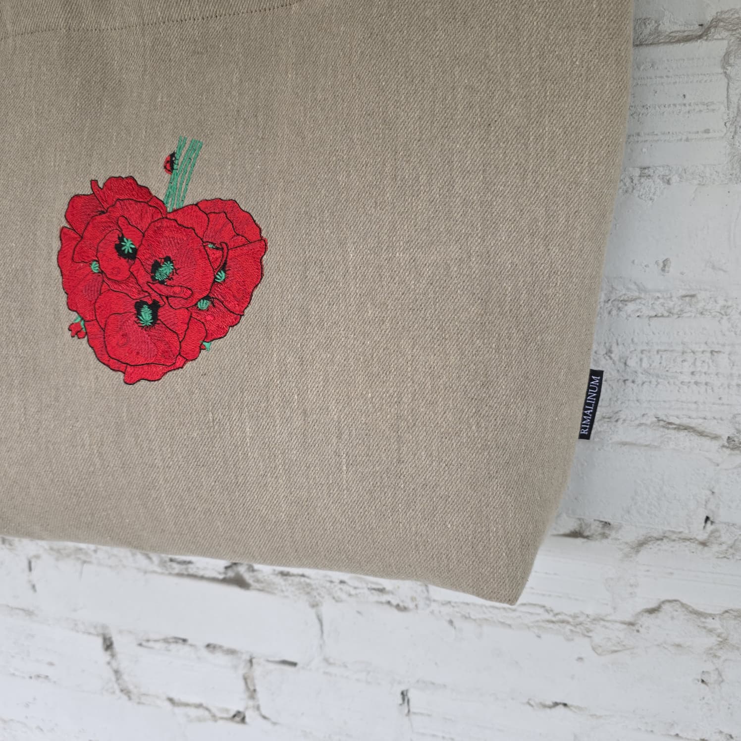 A beige linen tote bag with a red poppy cluster embroidered on the front, shown against a white brick wall background.