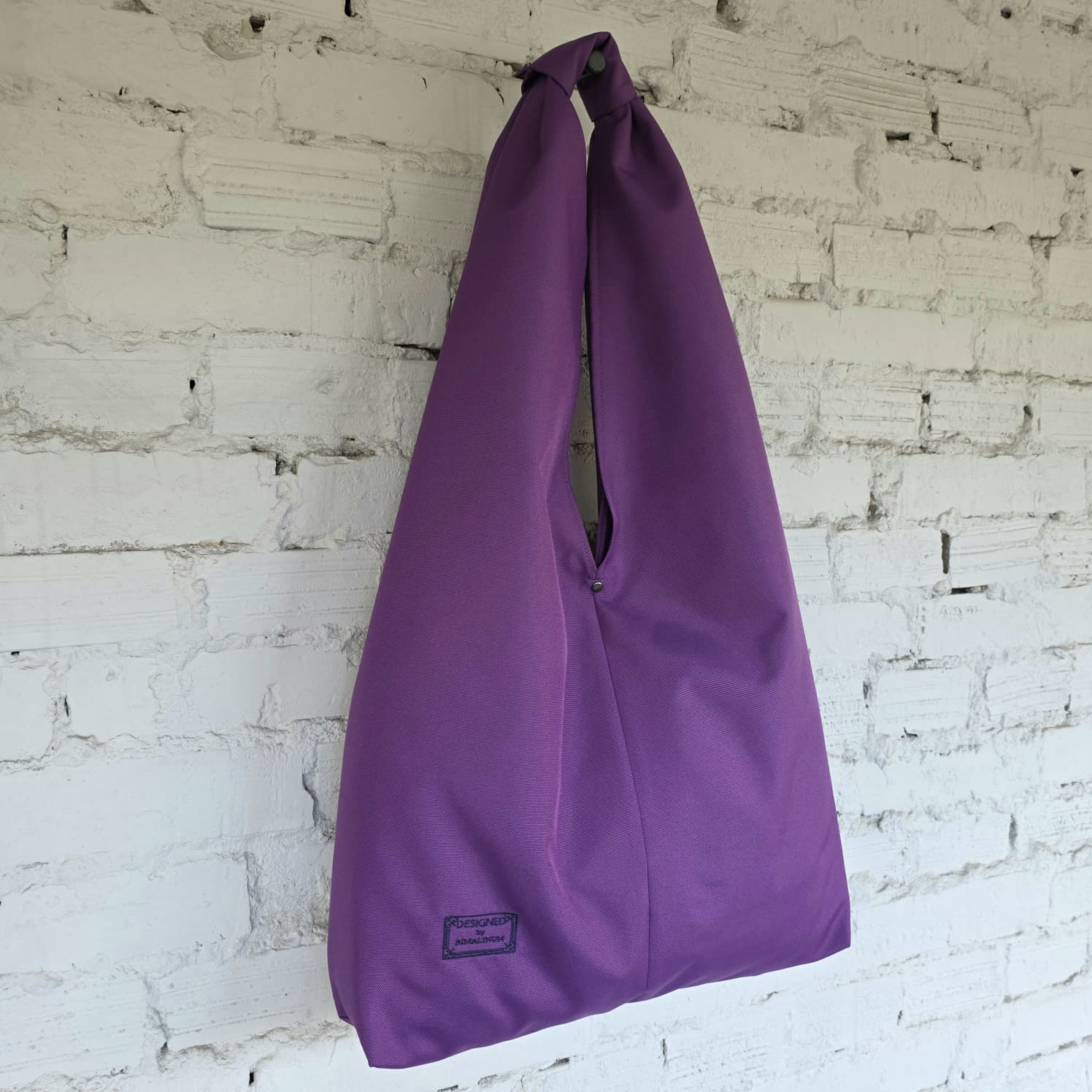 Oversized purple hobo shoulder bag hanging against a white brick wall, showing its soft slouchy silhouette.