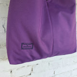 A close-up of a purple fabric bag or pouch with a small embroidered label reading 'DESIGNED by RIMALINUM', shown against a white painted brick wall.