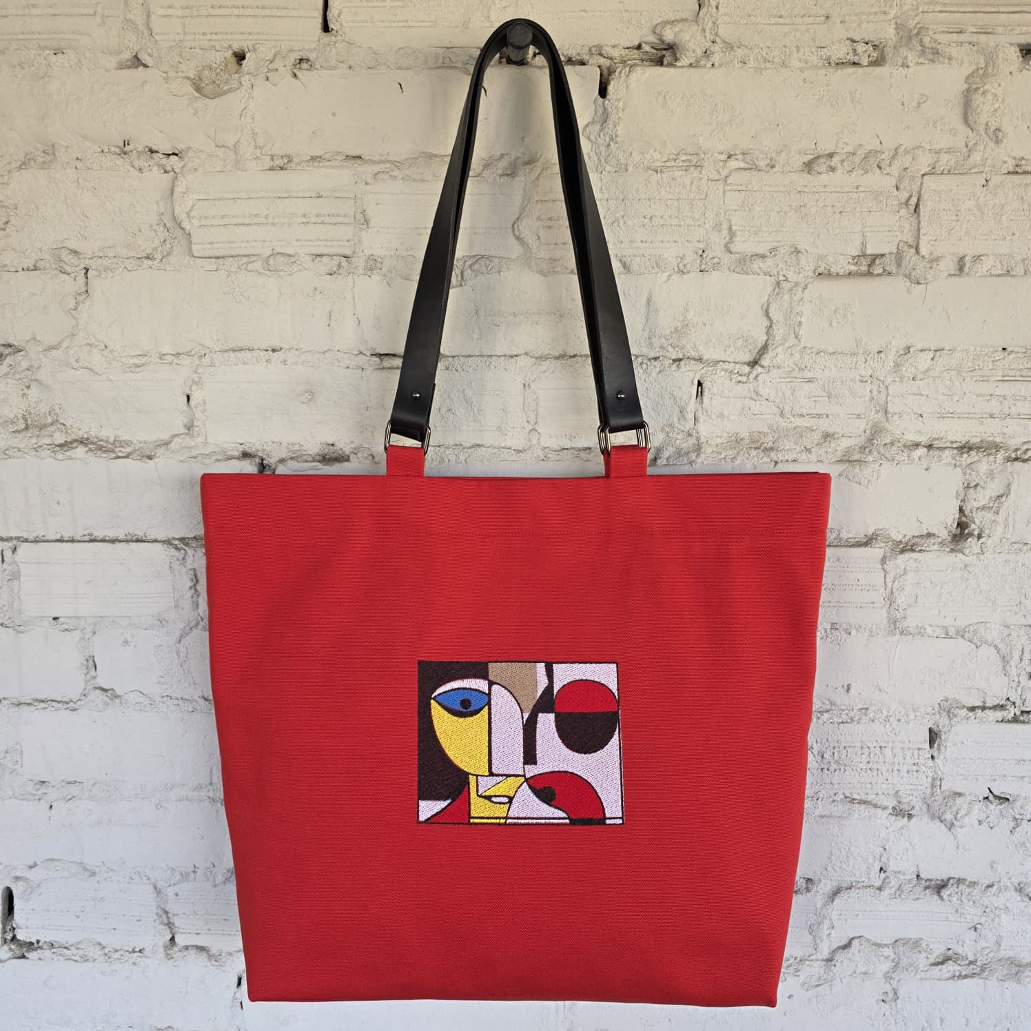 A red canvas tote bag with black shoulder straps, featuring a square abstract art embroidery on the front, hanging against a white brick wall.