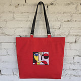 A red canvas tote bag with black shoulder straps, featuring a square abstract art embroidery on the front, hanging against a white brick wall.