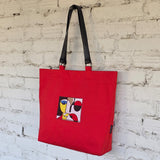 A red canvas tote bag with black shoulder straps hangs against a white painted brick wall. The front of the bag features a square abstract face artwork patch in black, red, yellow, white, and blue.