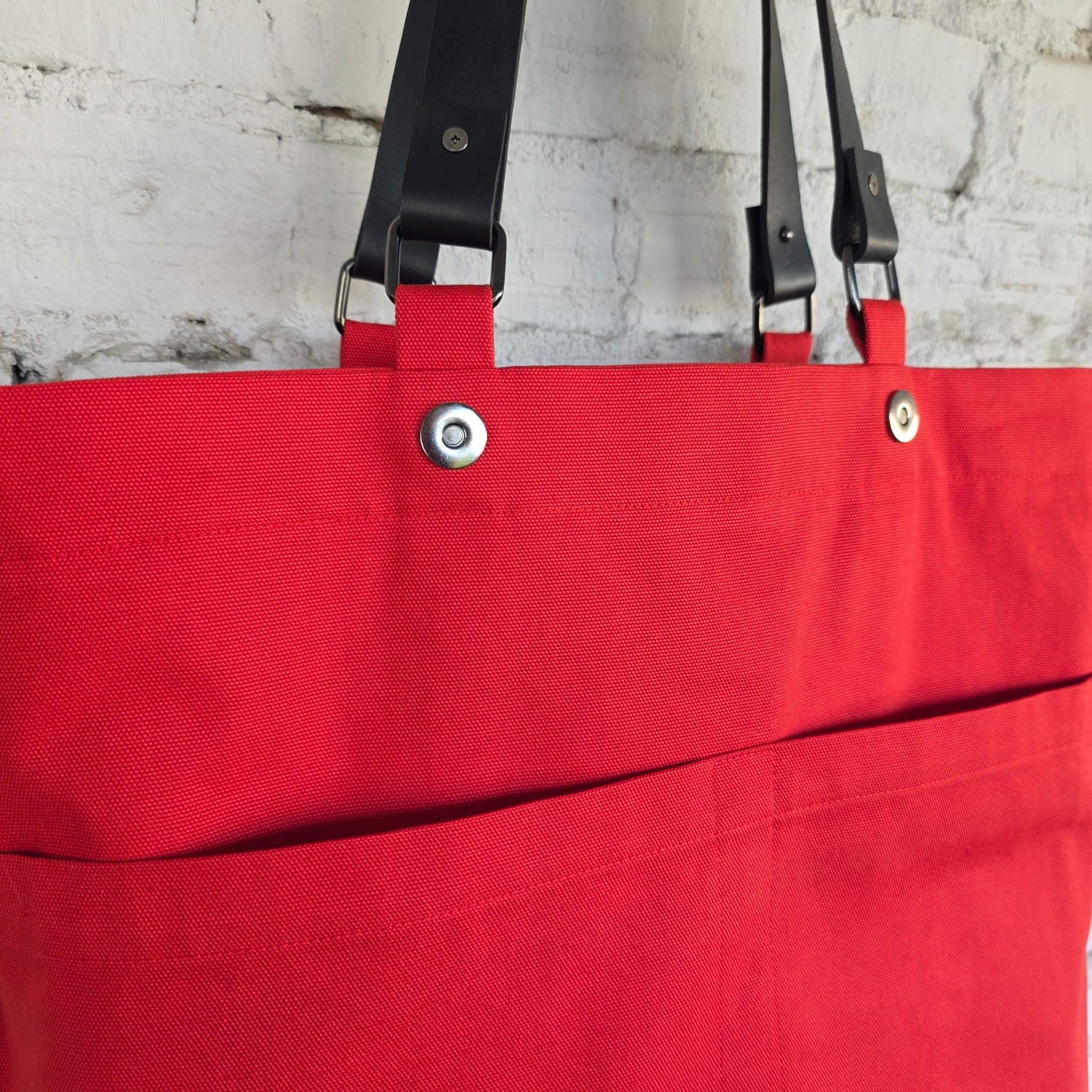 A close-up of a bright red canvas tote bag with black leather shoulder straps and silver metal snap buttons, hanging against a white brick wall.