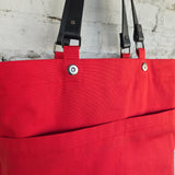 A close-up of a bright red canvas tote bag with black leather shoulder straps and silver metal snap buttons, hanging against a white brick wall.