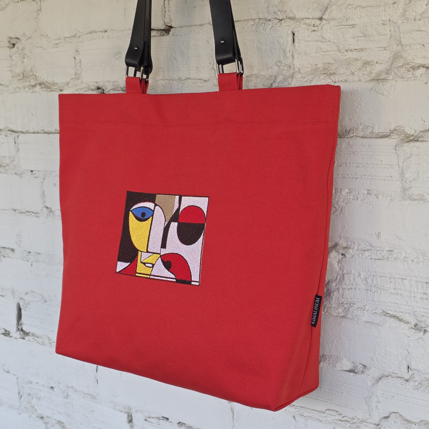 A red canvas tote bag with black shoulder straps, featuring a square abstract embroidered design on the front and a small black brand tag near the bottom right corner, hanging against a white brick wall