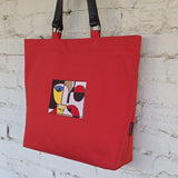 A red canvas tote bag with black shoulder straps, featuring a square abstract embroidered design on the front and a small black brand tag near the bottom right corner, hanging against a white brick wall