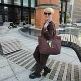 A woman with short blonde hair and sunglasses stands outdoors in a modern plaza, wearing a brown outfit and carrying a dark brown canvas tote bag .