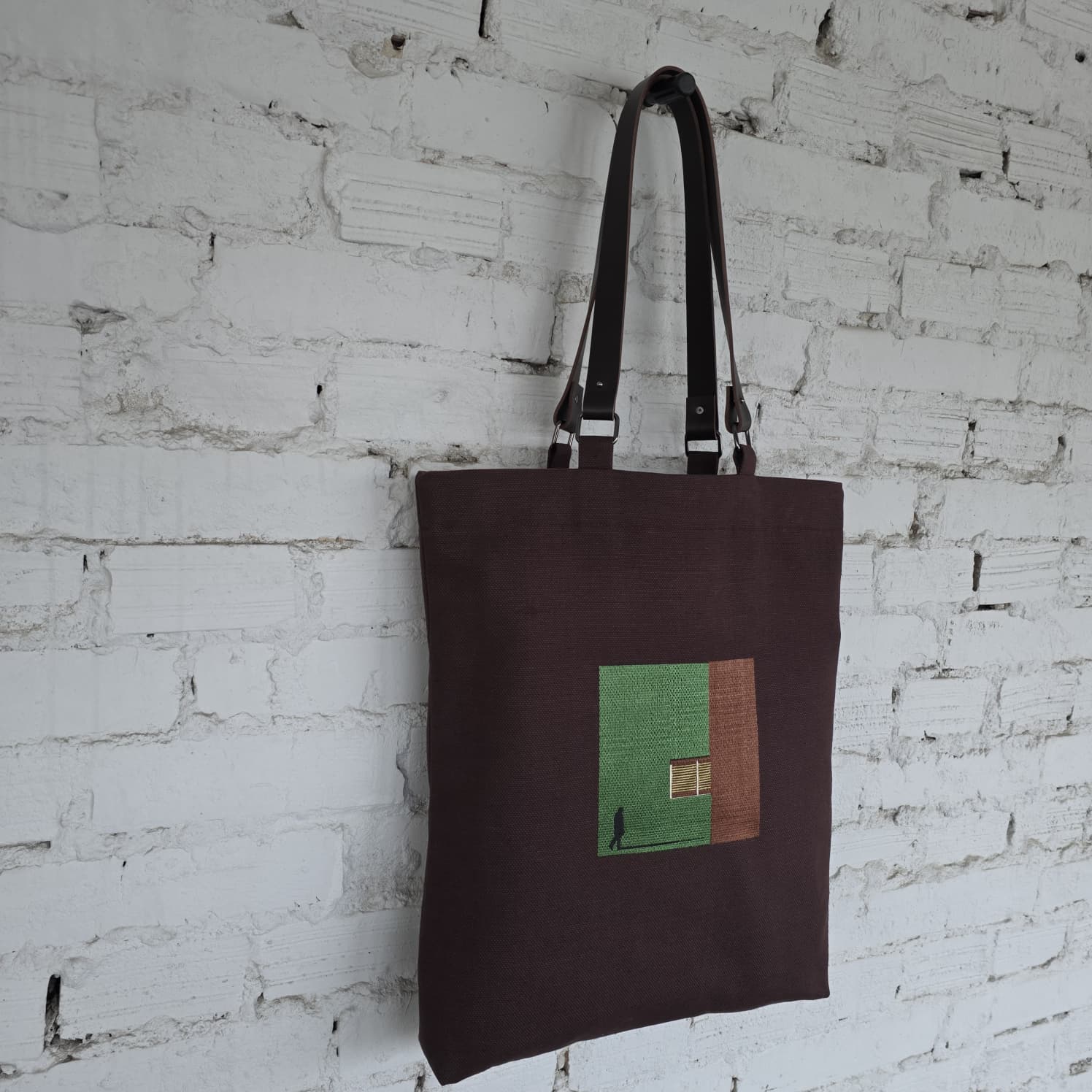 Brown tote bag with a green and brown square design against a white brick wall.